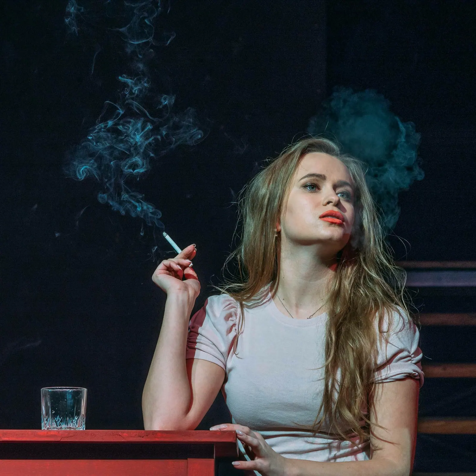 Krystyna on stage at a red table, holding a cigarette under blue light